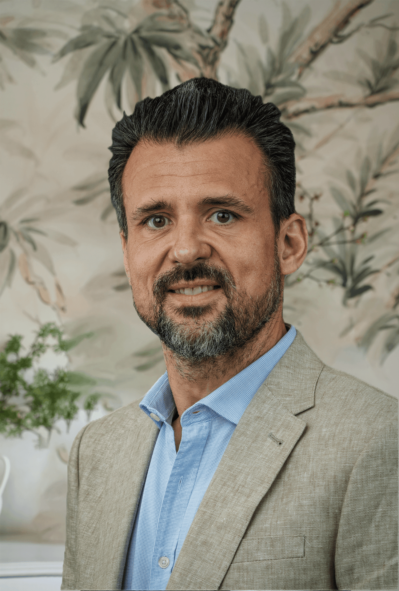 Man in a beige suit and blue shirt smiling with a leafy background.