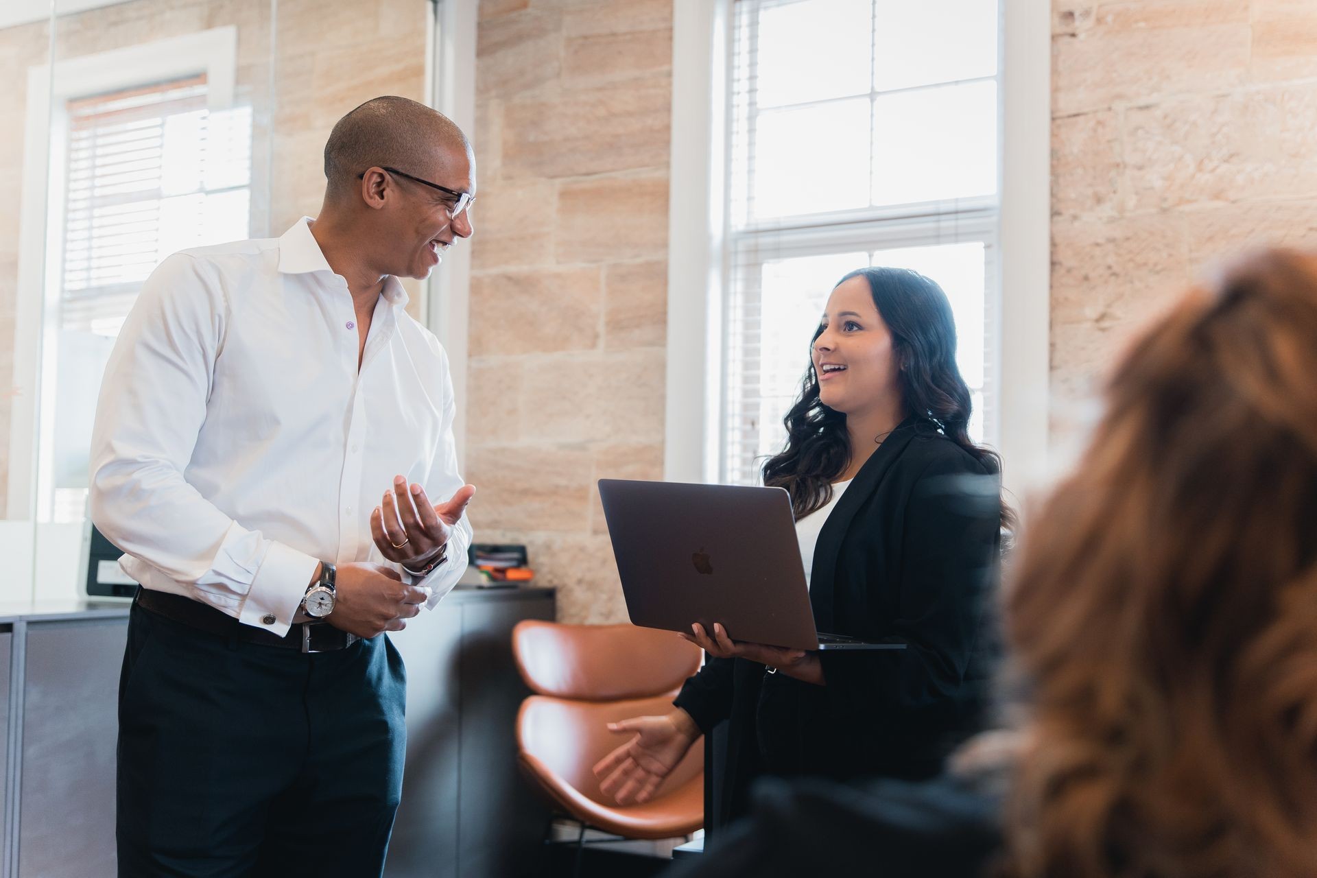 Two colleagues engage in conversation, with one holding a laptop, in a professional office setting.